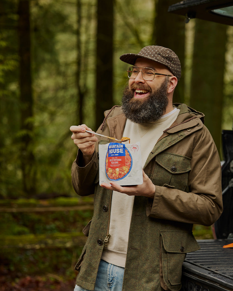 Man with a beard eating from a Mountain House pouch in front of an open truck in a forest