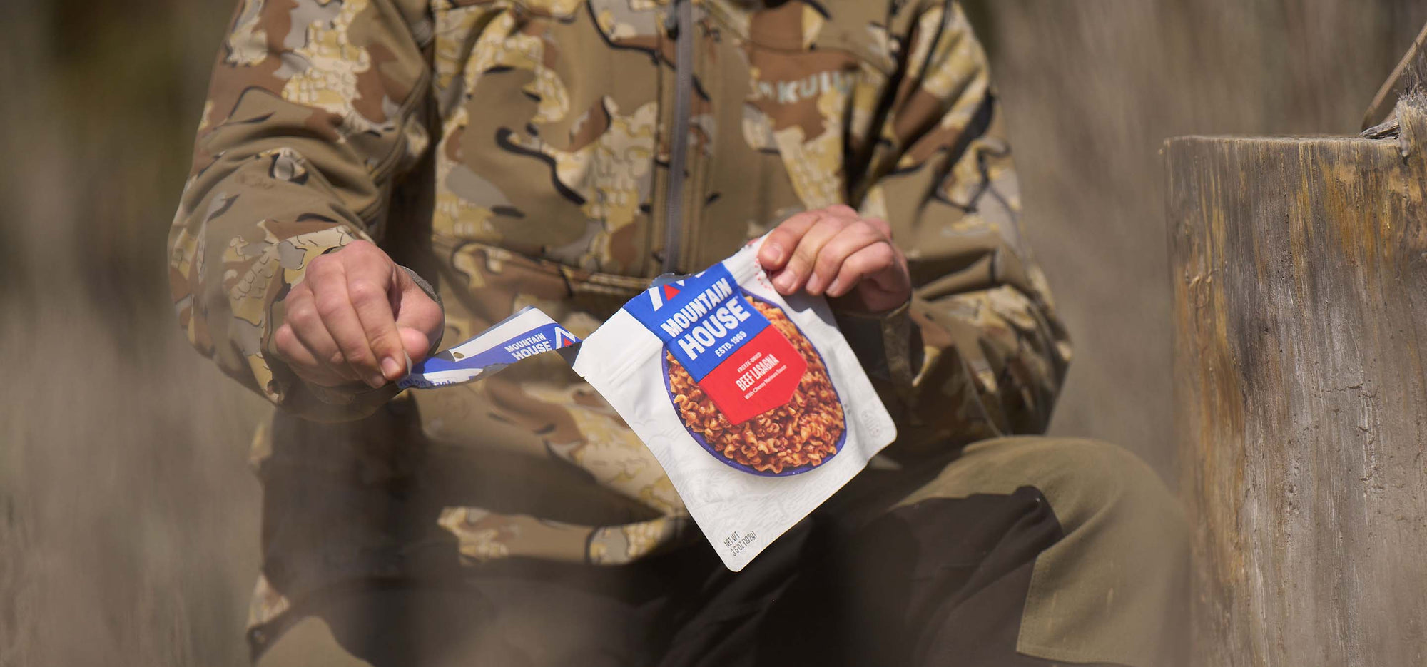 Person knelt down in camouflage tearing open pouch of Mountain House Beef Stew out in a clear cut.