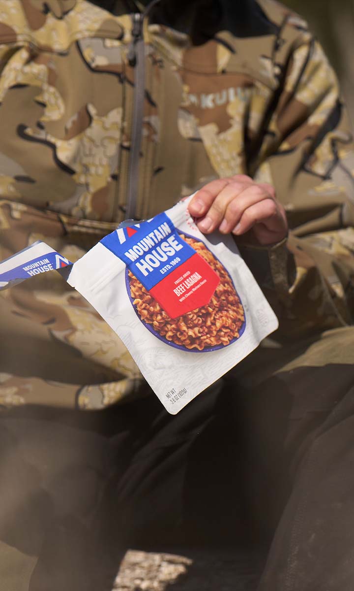 Person knelt down in camouflage tearing open pouch of Mountain House Beef Stew out in a clear cut.