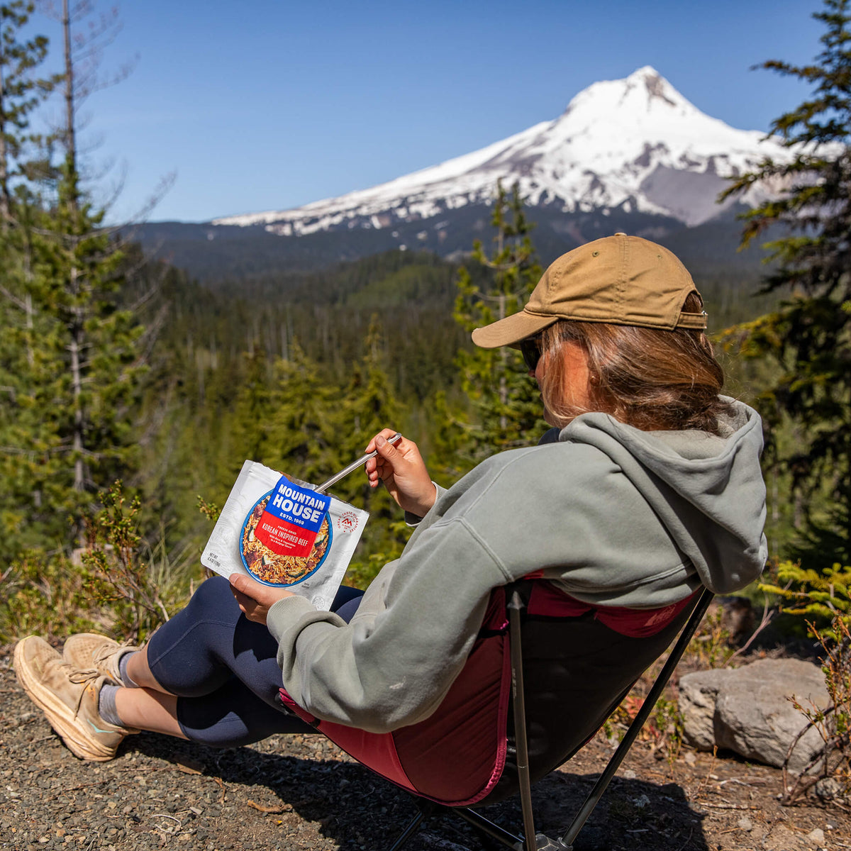 Girl seated in a camp chair eating from a Korean-Inspired Beef pouch with a snow-covered mountain in the background.