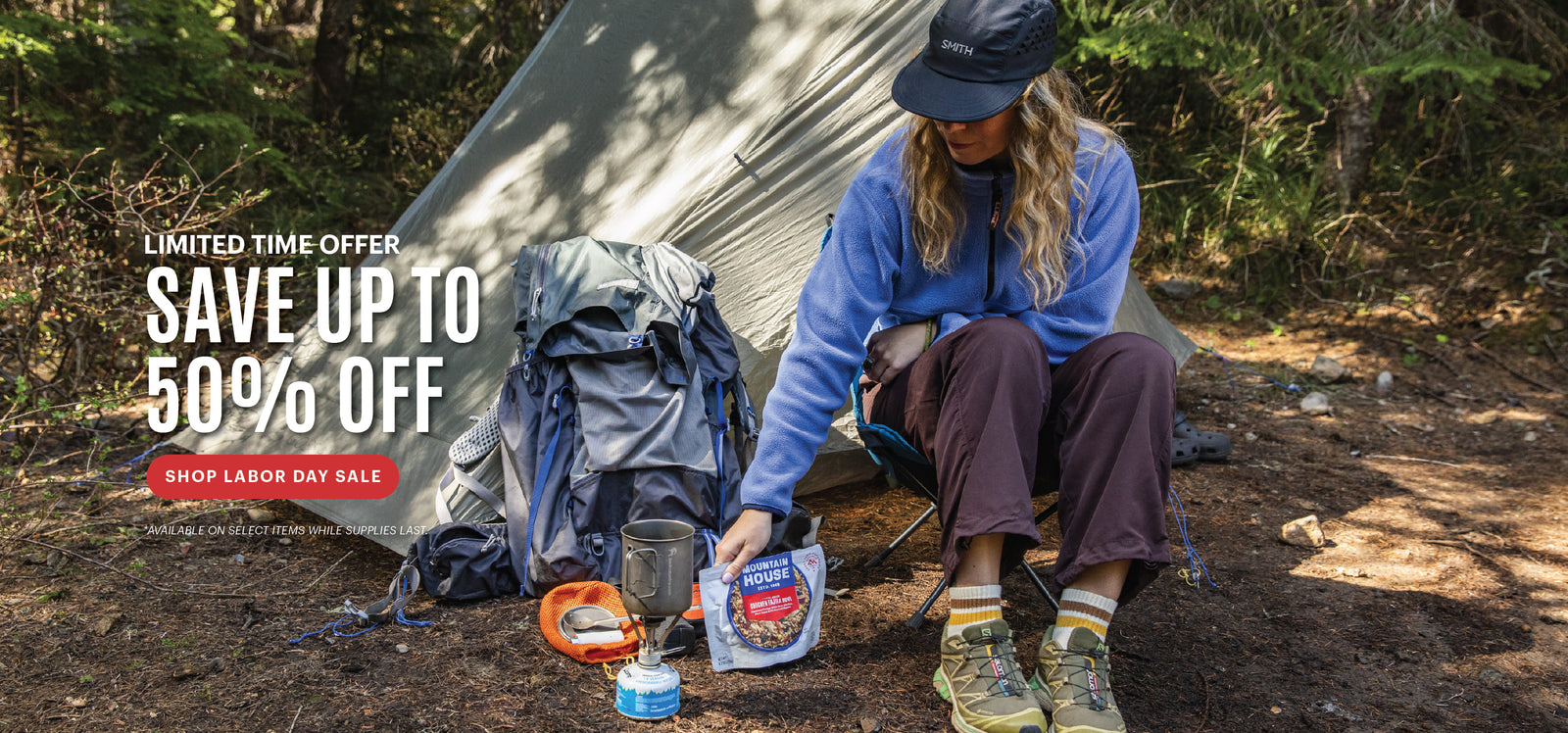 Woman reaching for a Mountain House pouch by a tent with Labor Day savings messaging of up to 50% off.