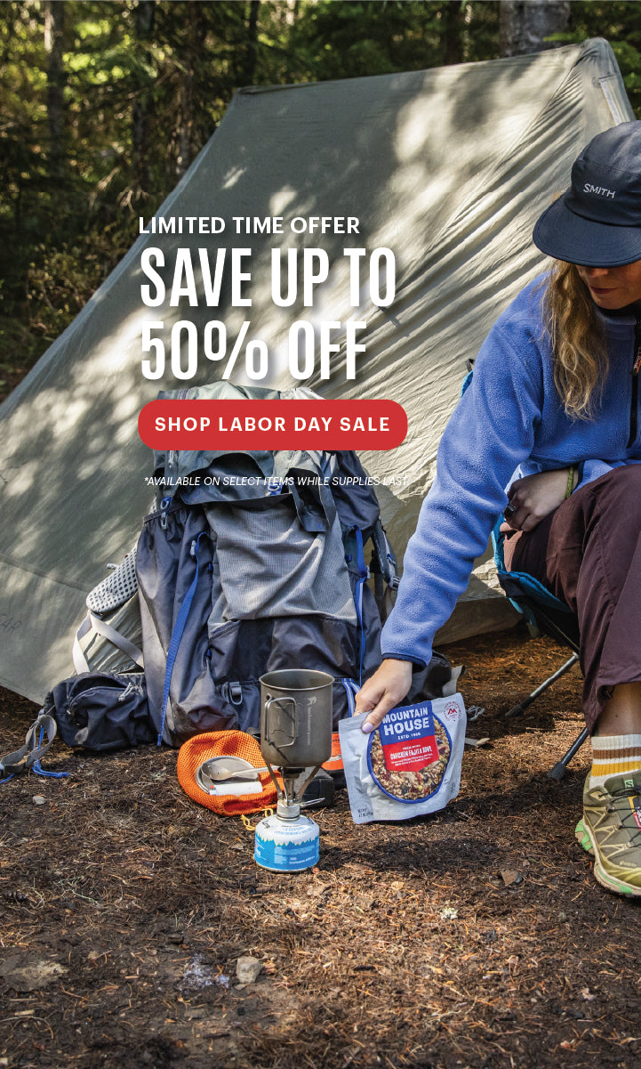 Woman reaching for a Mountain House pouch by a tent with Labor Day savings messaging of up to 50% off.