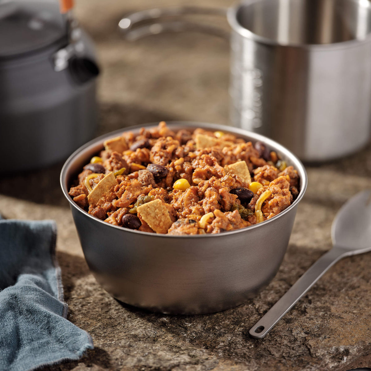 Metal bowl of beef enchilada with tortillas, beans, corn, and rice on a countertop with camping cookware in the background.