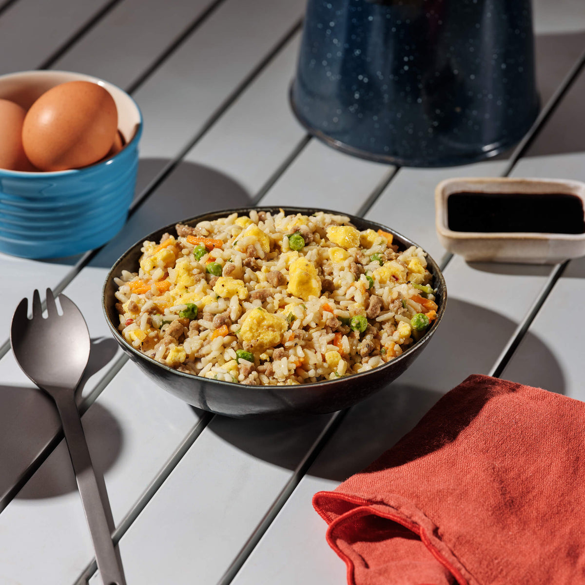 Bowl of breakfast fried rice with vegetables on a wooden table with eggs and a container of sauce in the background.
