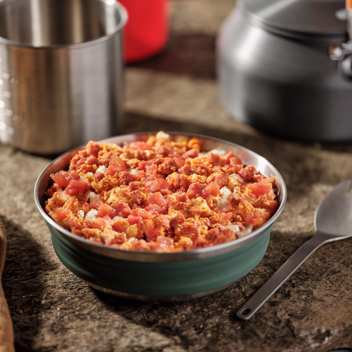 Bowl of cheesy pepperoni pizza on a rock surface with a camping kettle and cup in the background.