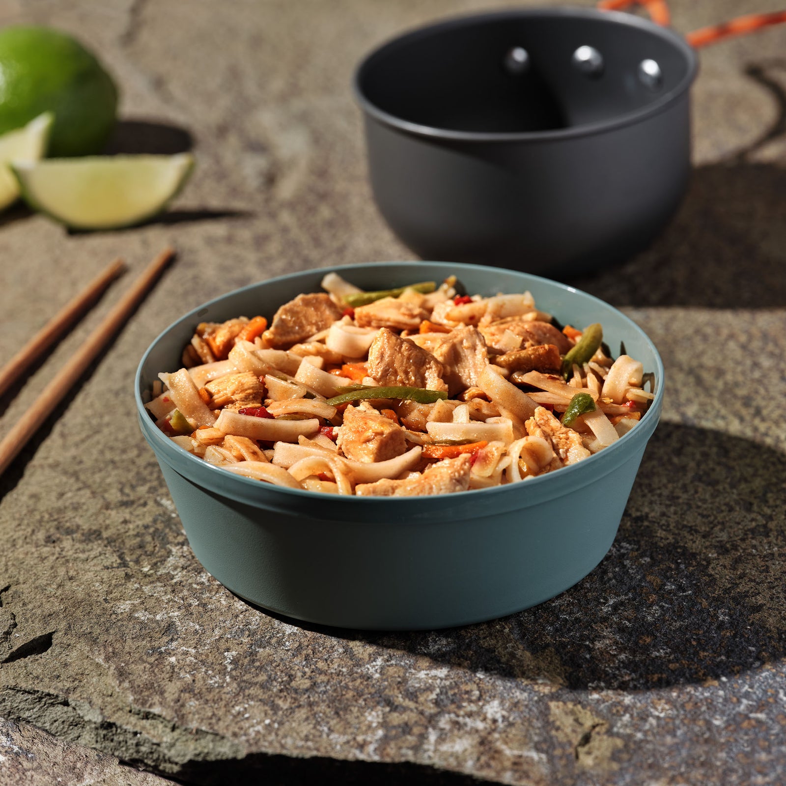 A blue camping bowl filled with Mountain House Pad Thai with Chicken rests on a rock surface with camping gear and limes in the background.