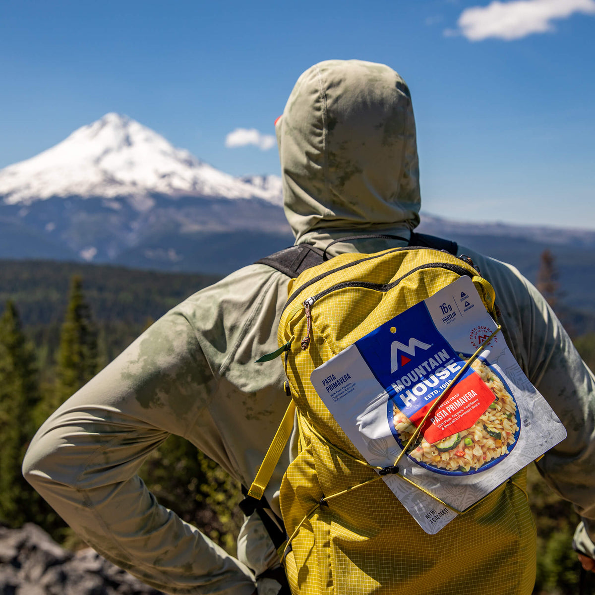Hiker with a yellow backpack and attached Pasta Primavera pouch, looking at a snow-capped mountain peak.