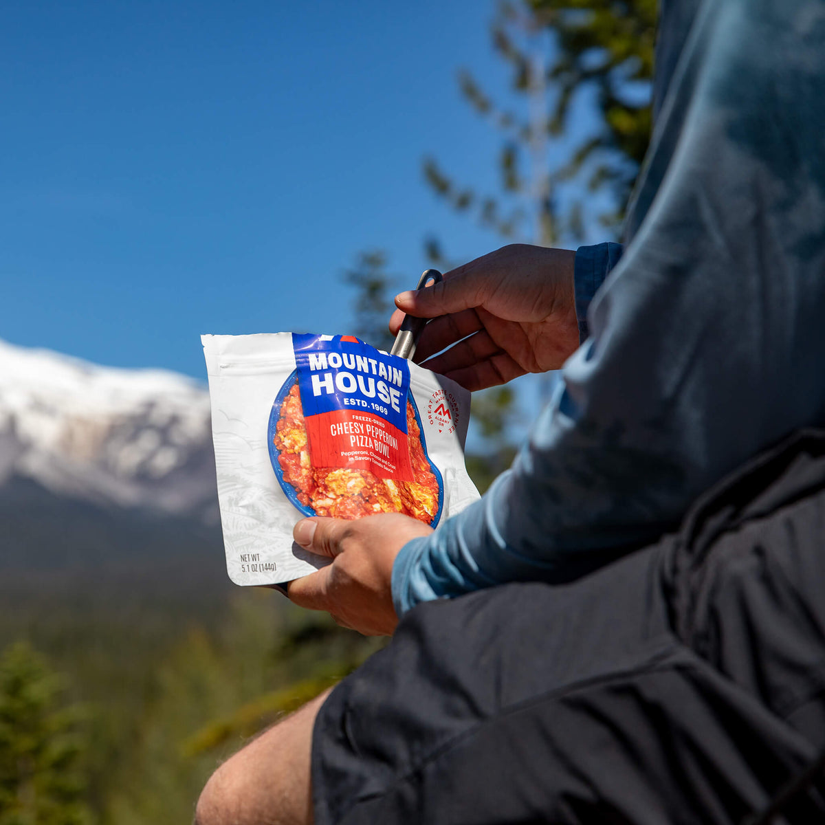 Person sitting down holding a spork in a Mountain House Pizza Bowl pouch in the outdoors.