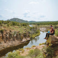 Person sitting on a rock overlooking a river with mountains in the background eating from a Mountain House Pro-Pak