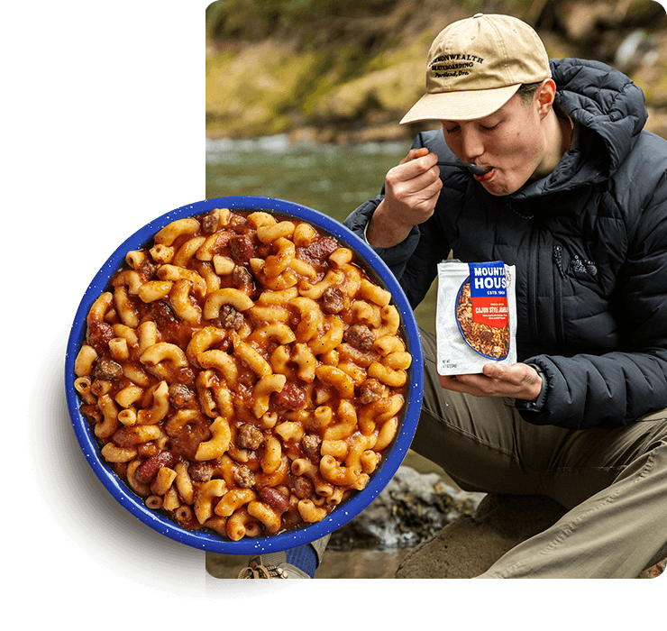 Person enjoying a Mountain House Chili Mac meal beside a river while sitting on a rock, with a bowl of Chili Mac in the foreground.