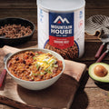 Mountain House Freeze-Dried Ground Beef on a wood countertop with a bowl of chili, avocado, and utensils.